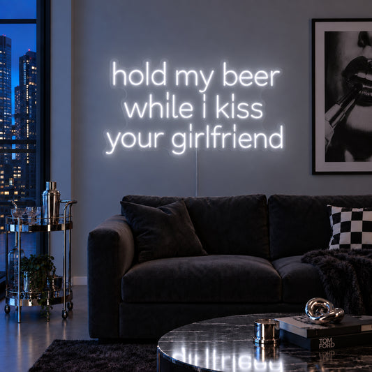 Hold My Beer While I Kiss Your Girlfriend Neon Sign glowing white above a burgundy sofa.