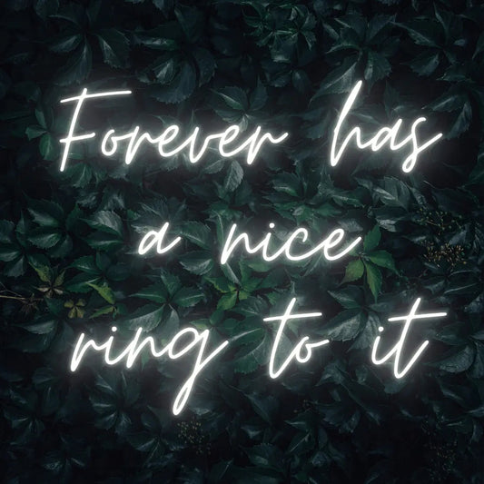 Forever Has a Nice Ring to It LED Neon Light Sign in white, featuring three lines of script lettering against a dark foliage backdrop.