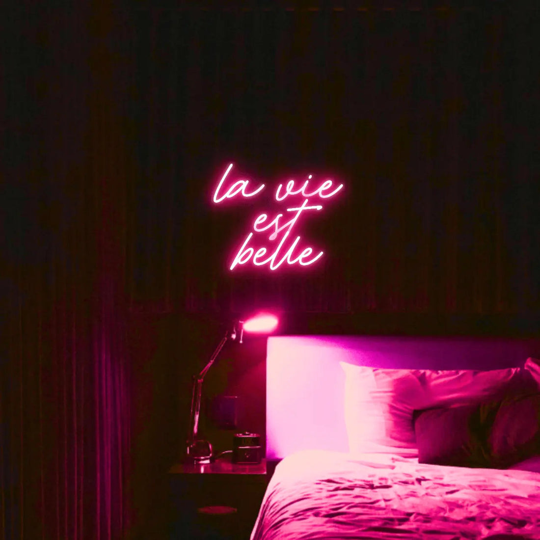 La Vie est Belle LED Neon Light Sign in pink, featuring script lettering illuminated above a bed.