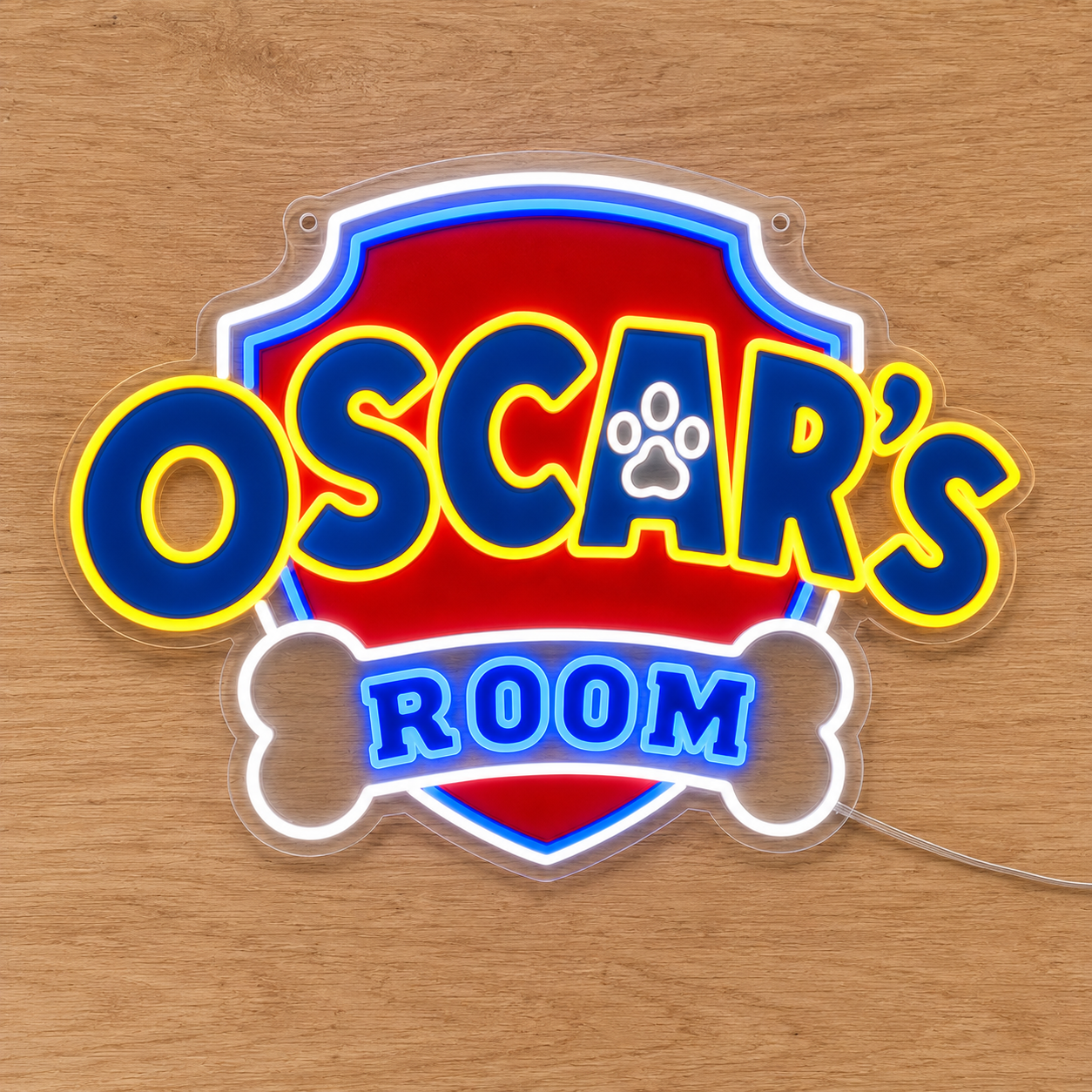 A custom Paw Patrol acrylic LED neon-style sign reading 'OSCAR'S ROOM' with a red shield background, blue lettering outlined in yellow, a white bone shape, and a paw print inside the letter A, displayed on a light wood surface.