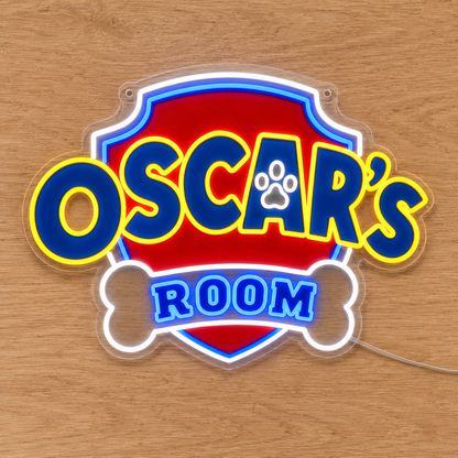 A custom Paw Patrol acrylic LED neon-style sign reading 'OSCAR'S ROOM' with a red shield background, blue lettering outlined in yellow, a white bone shape, and a paw print inside the letter A, displayed on a light wood surface.
