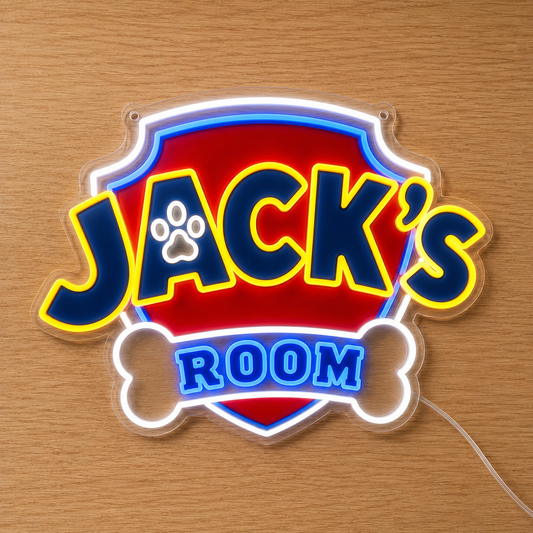 A custom Paw Patrol LED neon-style sign shaped like a badge with a bone outline, reading "Jack's Room" in blue letters with yellow and white glowing borders, placed on a light wood surface.