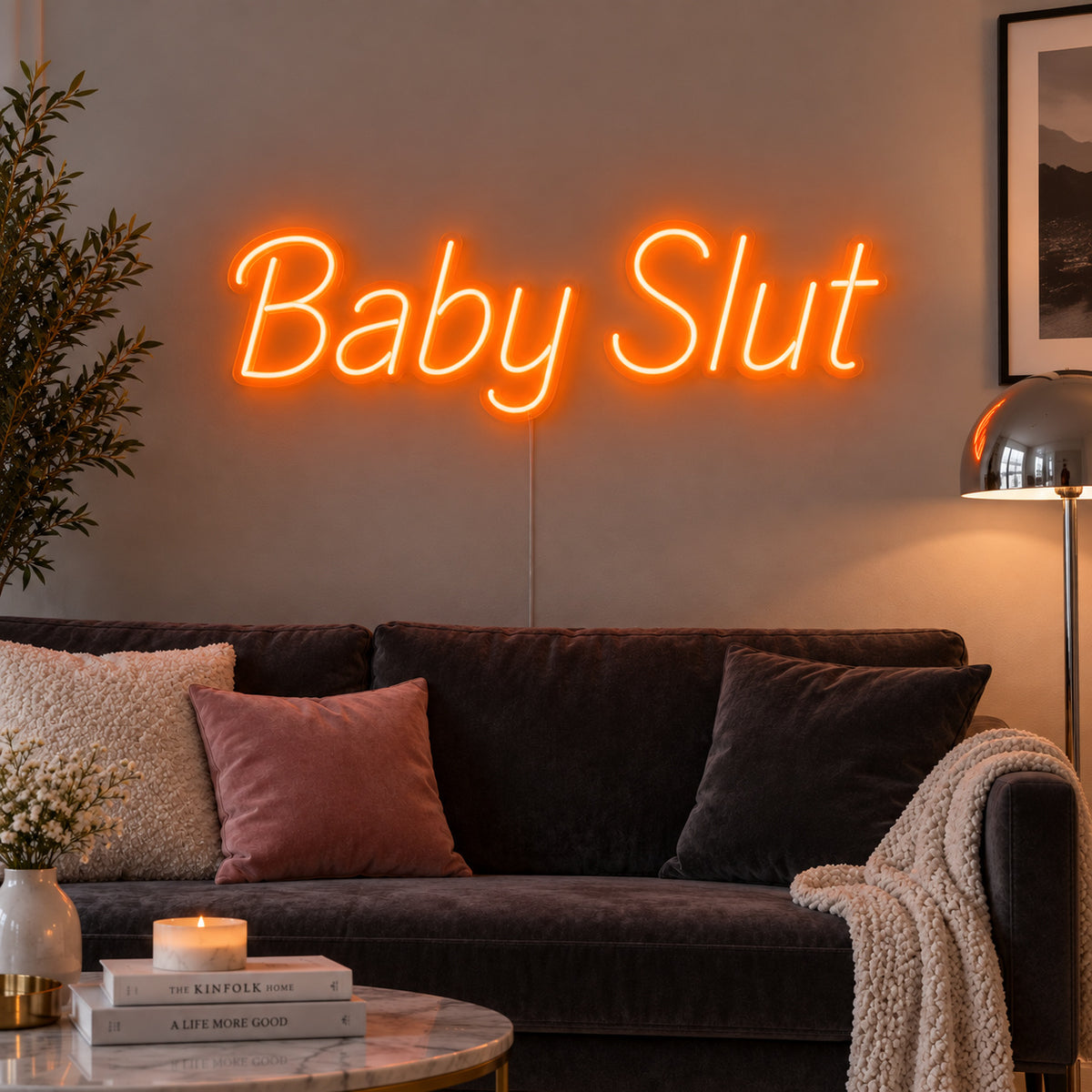 Orange version of the Baby Slut Neon Sign, shown above a grey sofa with pink cushions.
