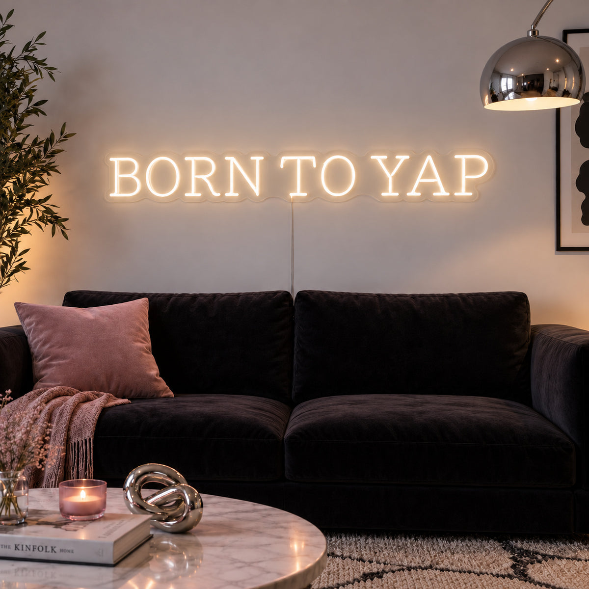 Warm White version of the Born To Yap Neon Sign, shown above a dark sofa and glass coffee table.