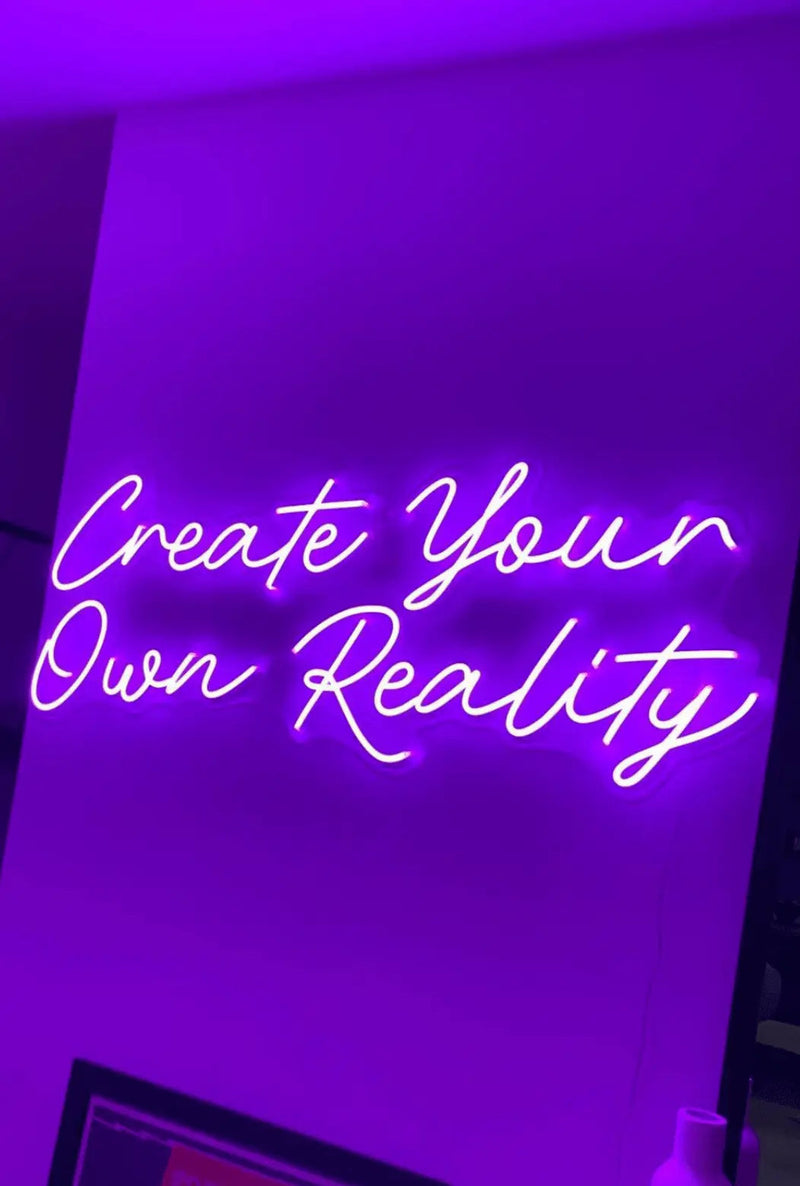 Create Your Own Reality LED Neon Light Sign in purple, featuring two lines of script lettering on a purple-lit wall.