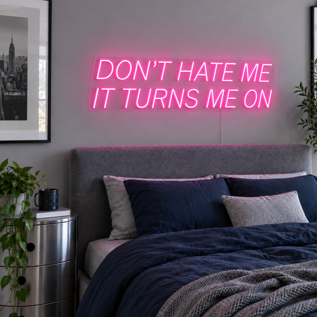 Don't Hate Me It Turns Me On Neon Sign glowing pink above a bed with dark blue bedding.