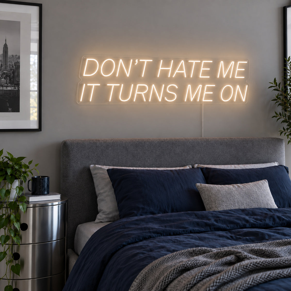 Warm White version of the Don't Hate Me It Turns Me On Neon Sign, shown above a bed with dark blue bedding.