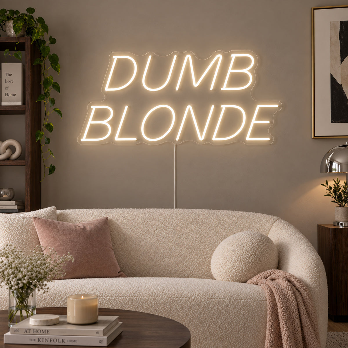 Warm White version of the Dumb Blonde Neon Sign, shown in slanted lettering above a cream sofa.