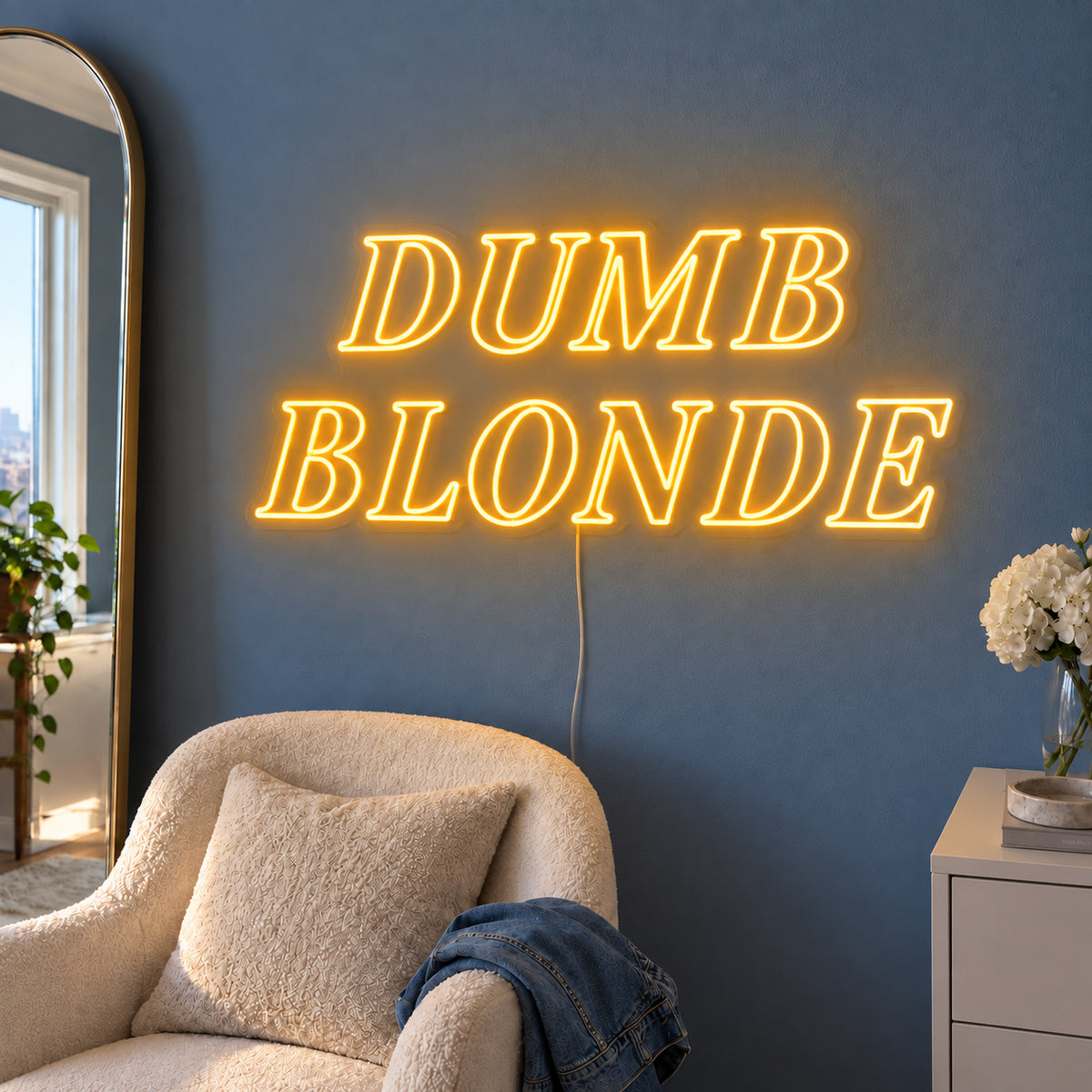 Dumb Blonde Neon Sign glowing gold in outlined serif lettering above a cream chair.