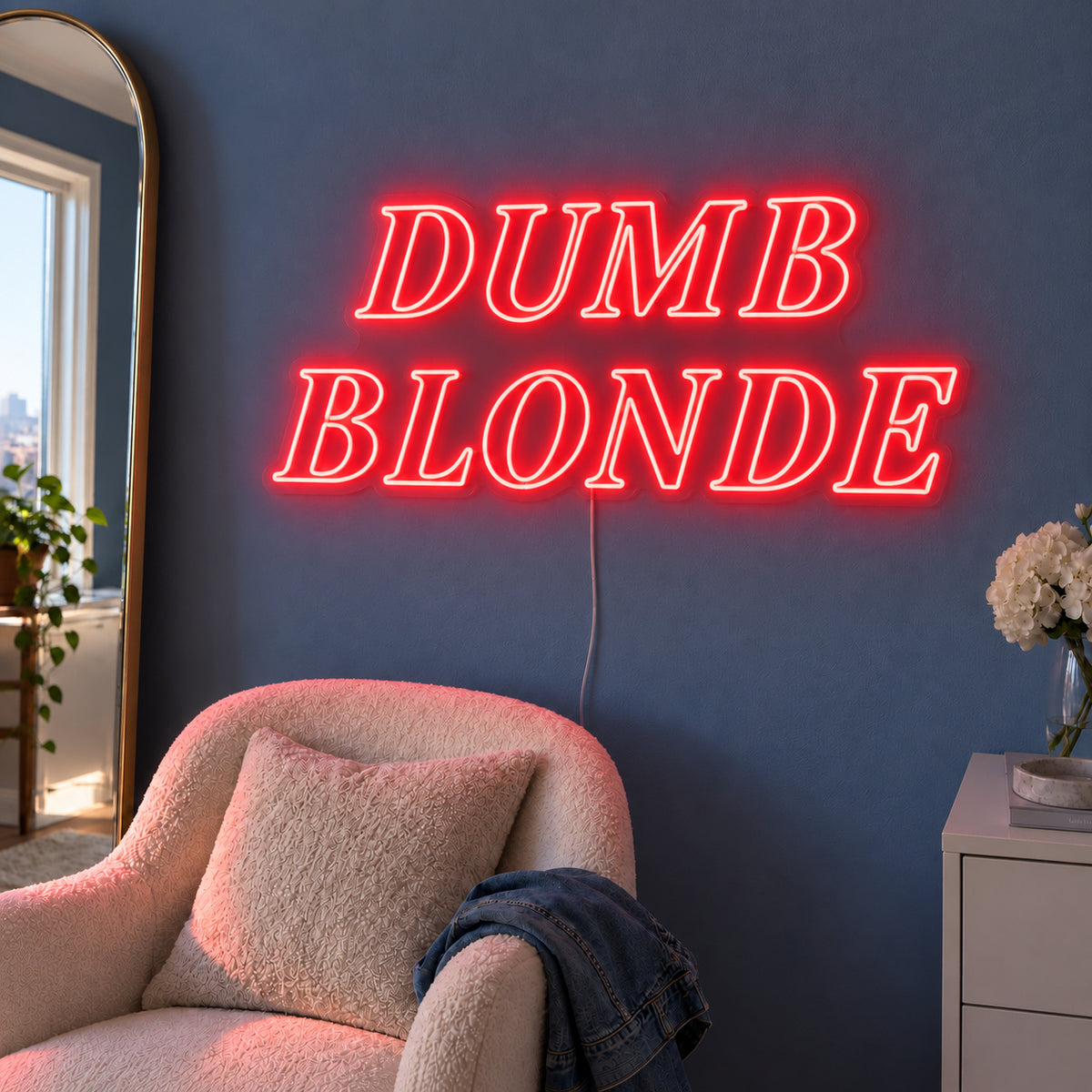 Dumb Blonde Neon Sign with red lighting, displayed in outlined serif lettering above a cream chair.