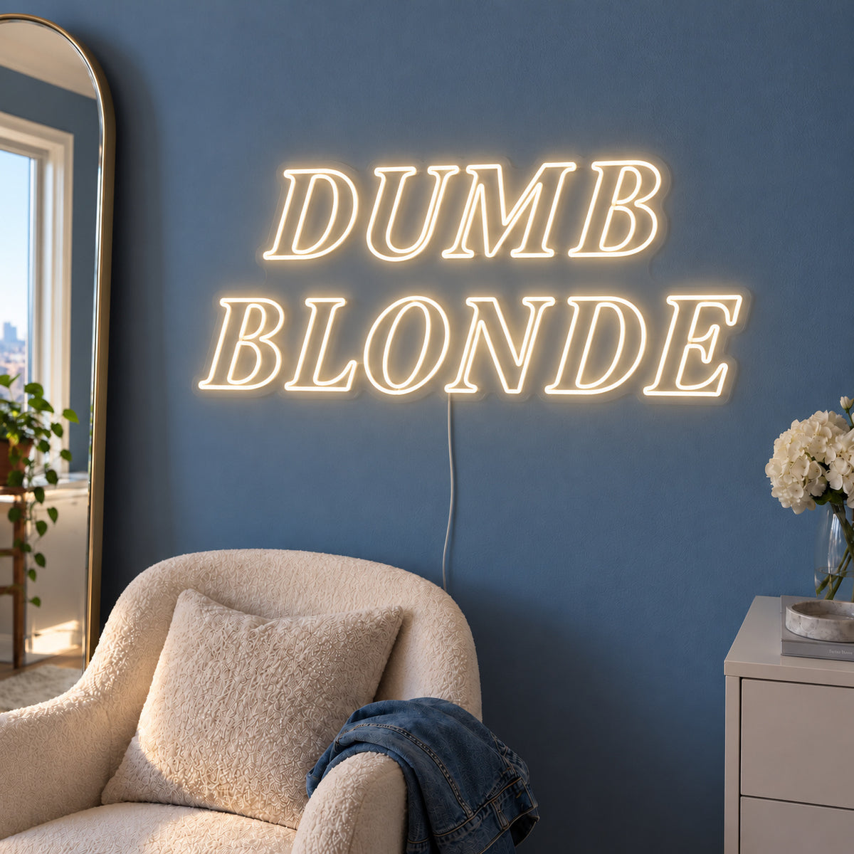 Warm White version of the Dumb Blonde Neon Sign, shown in outlined serif lettering above a cream chair.