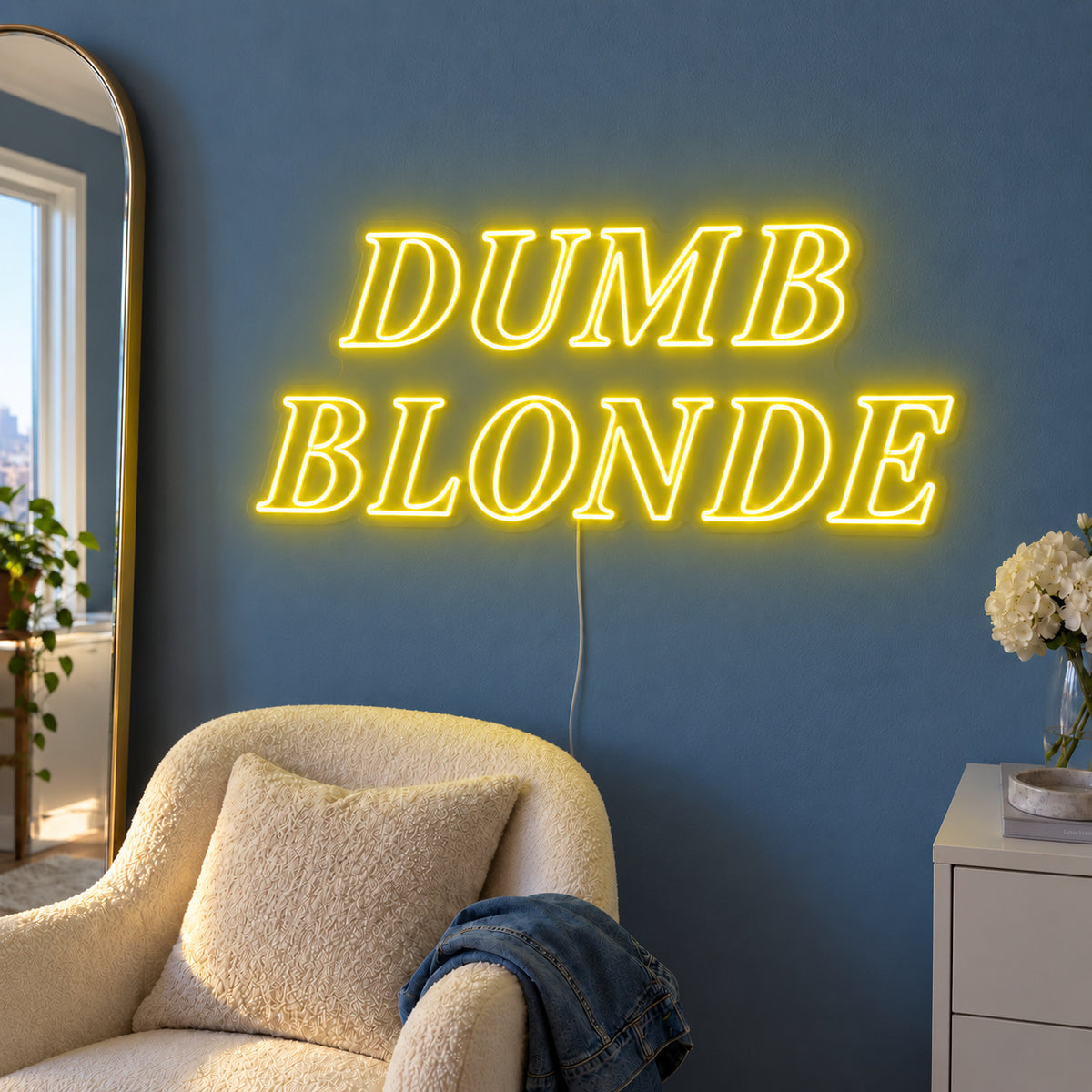 Dumb Blonde Neon Sign with yellow lighting, displayed in outlined serif lettering above a cream chair.