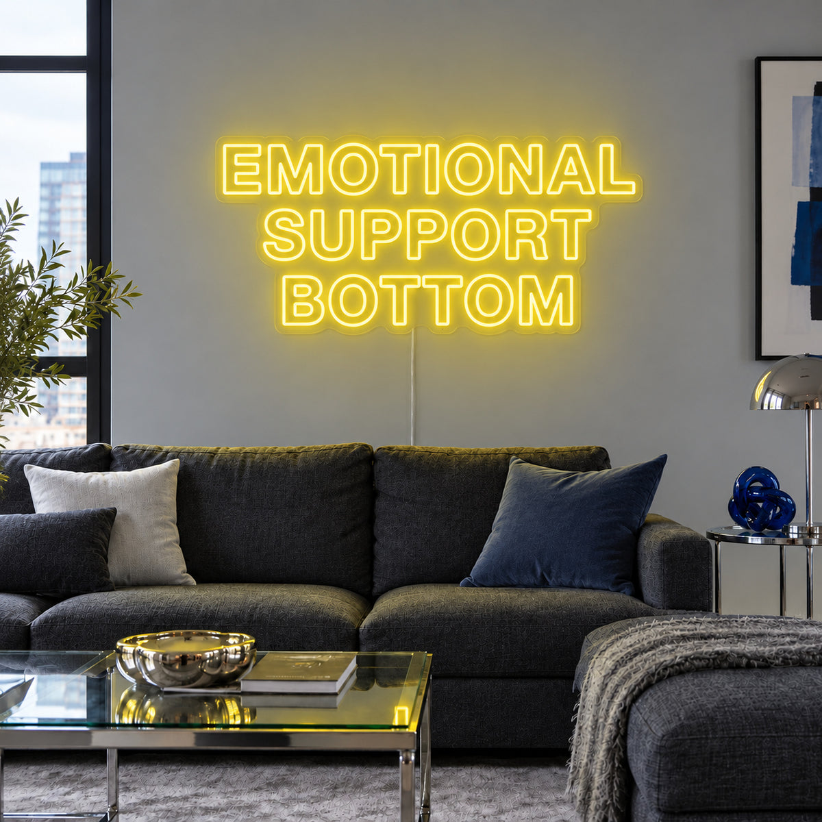 Emotional Support Bottom Neon Sign with yellow lighting, displayed above a charcoal sofa.
