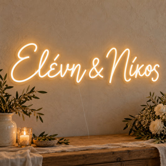 Personalised Greek Couple Names Neon Sign on clear cut-to-shape acrylic in an illuminated wall display; example personalisation shown.