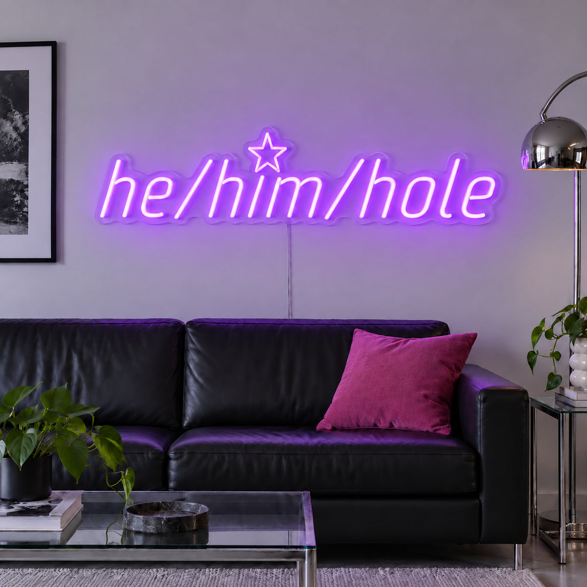 He/Him/Hole Neon Sign with purple lighting, displayed above a black leather sofa.