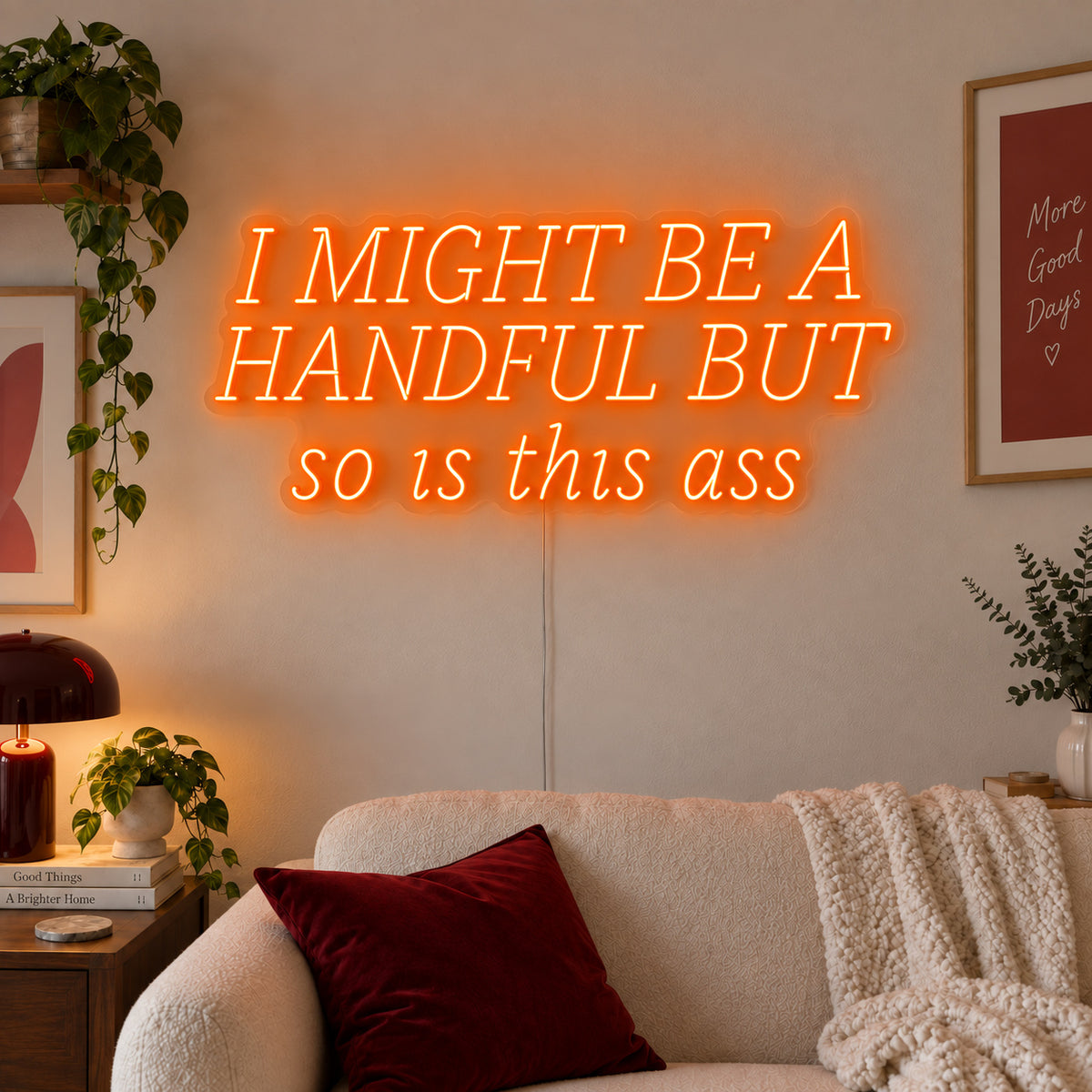 Orange version of the I Might Be A Handful But So Is This Ass Neon Sign, shown in wide lettering above a cream sofa.
