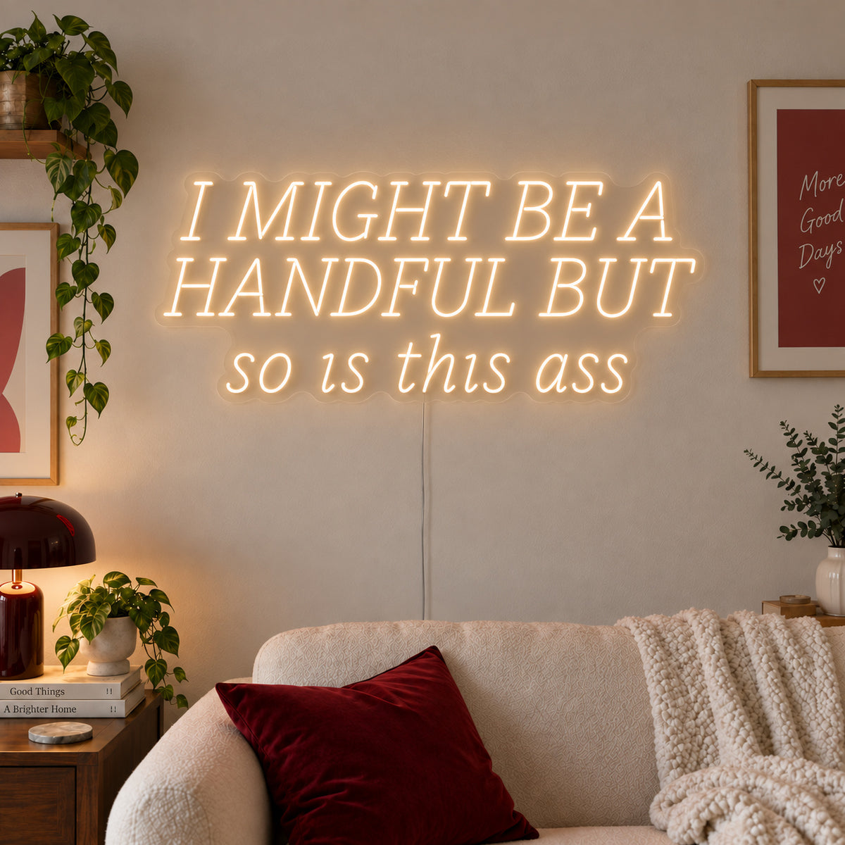 Warm White version of the I Might Be A Handful But So Is This Ass Neon Sign, shown in wide lettering above a cream sofa.