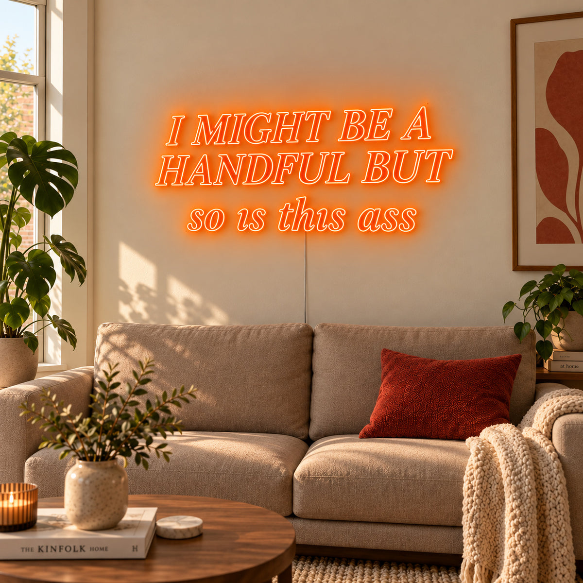 Orange version of the I Might Be A Handful But So Is This Ass Neon Sign, shown in compact lettering above a cream sofa.