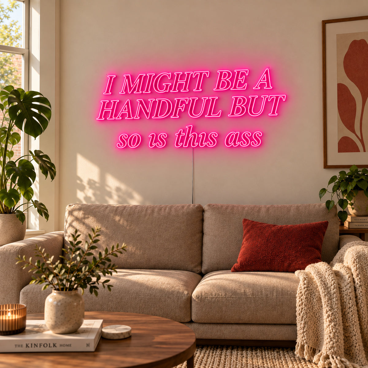 Rose Red version of the I Might Be A Handful But So Is This Ass Neon Sign, shown in compact lettering above a cream sofa.