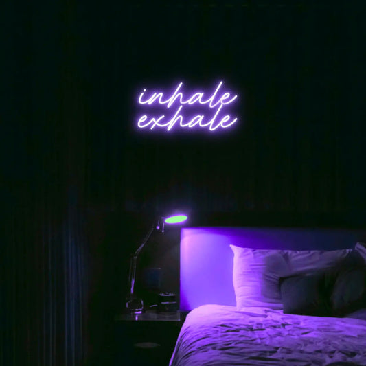 Inhale Exhale LED Neon Light Sign in purple, featuring two lines of script lettering illuminated above a bed.