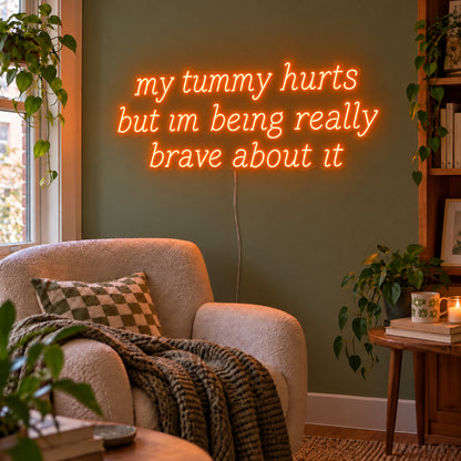 Orange version of the My Tummy Hurts But Im Being Really Brave About It Neon Sign, shown above a reading chair against a green wall.