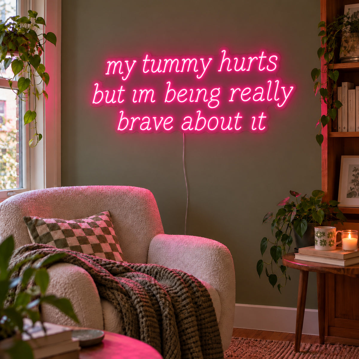 Rose Red version of the My Tummy Hurts But Im Being Really Brave About It Neon Sign, shown above a reading chair against a green wall.