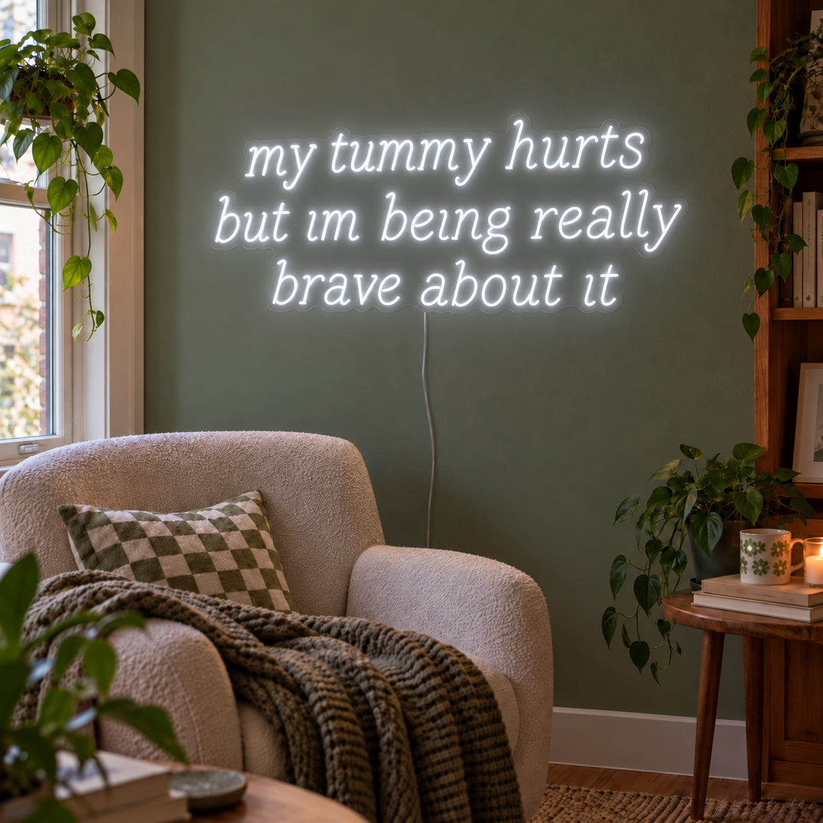 My Tummy Hurts But Im Being Really Brave About It Neon Sign glowing white above a reading chair against a green wall.