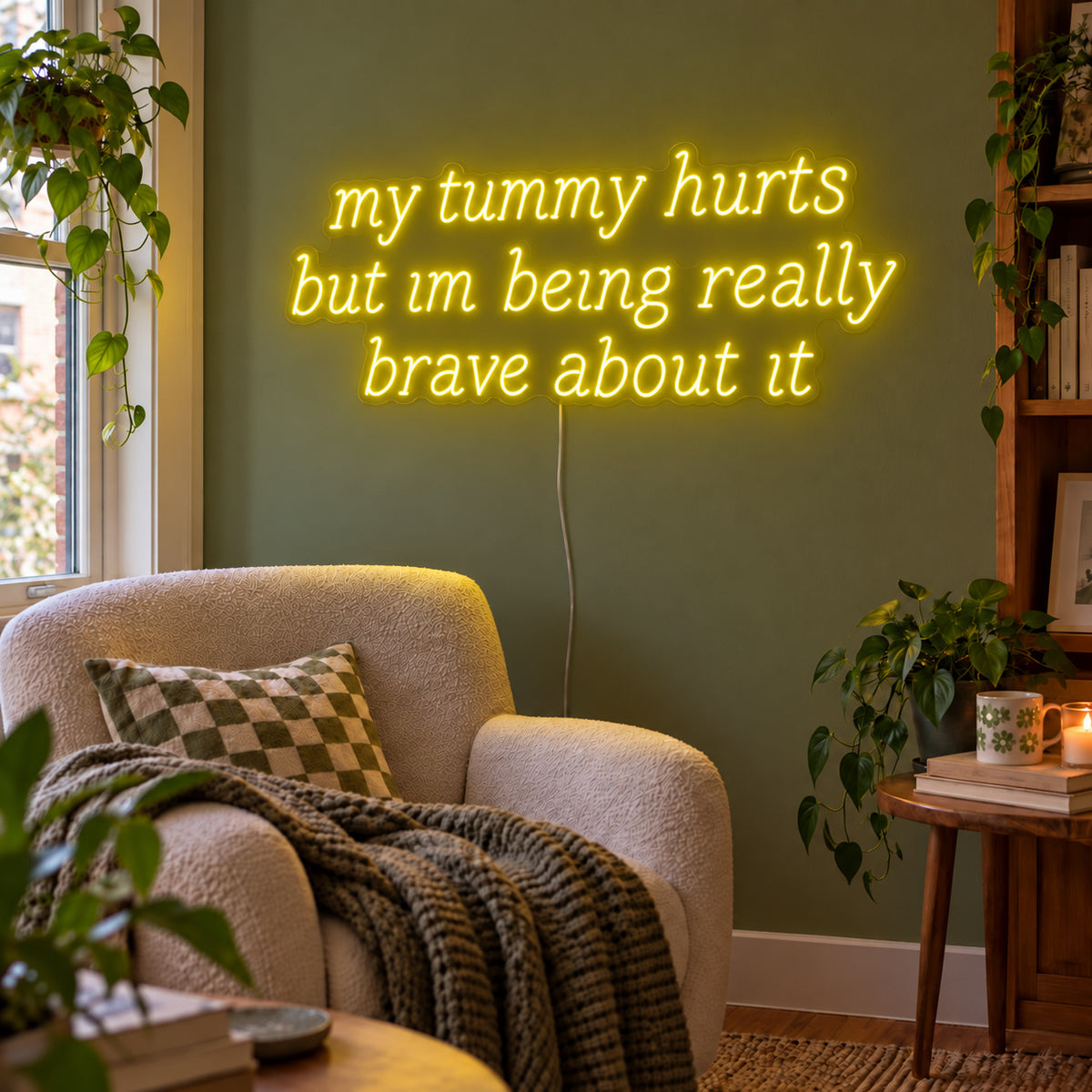 My Tummy Hurts But Im Being Really Brave About It Neon Sign with yellow lighting, displayed above a reading chair against a green wall.