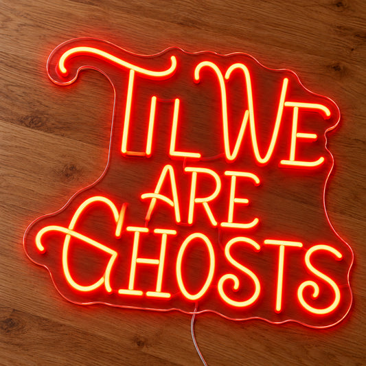 Til We Are Ghosts Ornate Neon Sign featuring Til We Are Ghosts wording in decorative orange lettering with curved flourishes, illuminated on clear acrylic lying on a natural oak wooden surface.